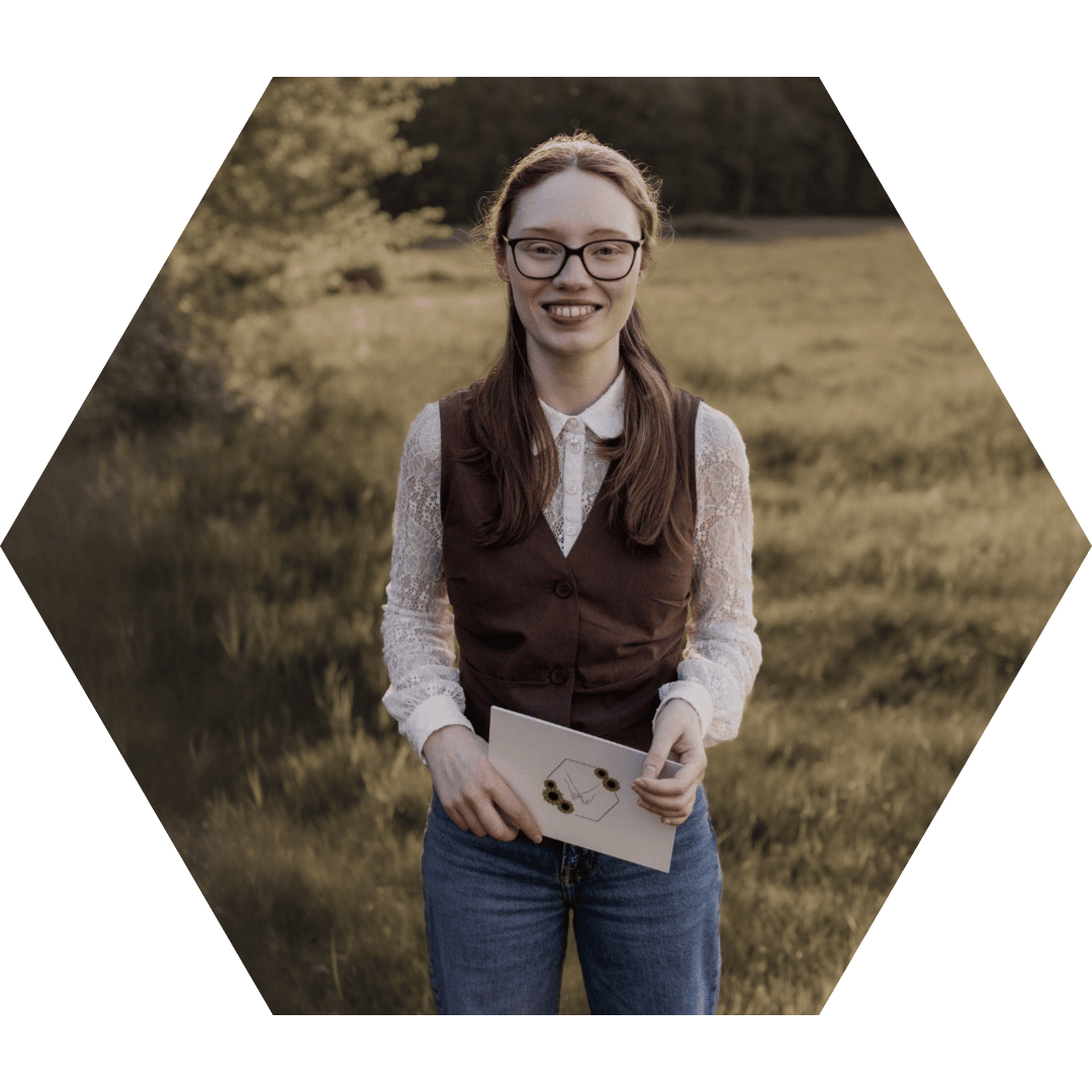 Samantha Liston steht lächelnd auf einer Wiese im Abendlicht. Sie trägt eine Spitzenbluse mit brauner Weste und hält eine Karte mit Sonnenblumenlogo in der Hand – als inklusive Traurednerin in NRW strahlt sie Offenheit und Wärme aus.